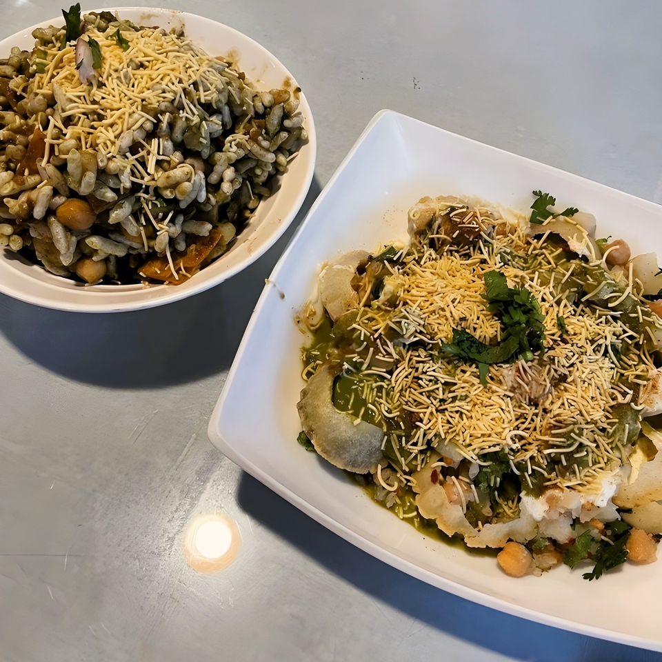 Two bowls of Indian chaat, one with puffed rice and the other with flatbread, topped with sauces and crispy noodles.