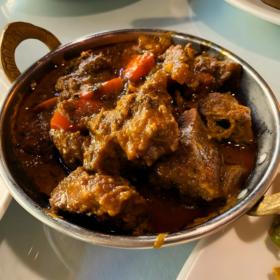 Close-up of a metal bowl filled with rich, dark Indian curry and chunks of meat and vegetables.