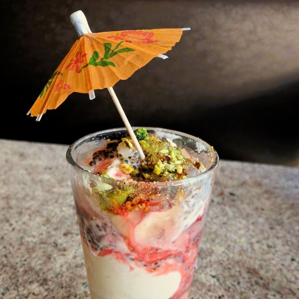 A glass of colorful Indian falooda with an orange cocktail umbrella, topped with pistachios.